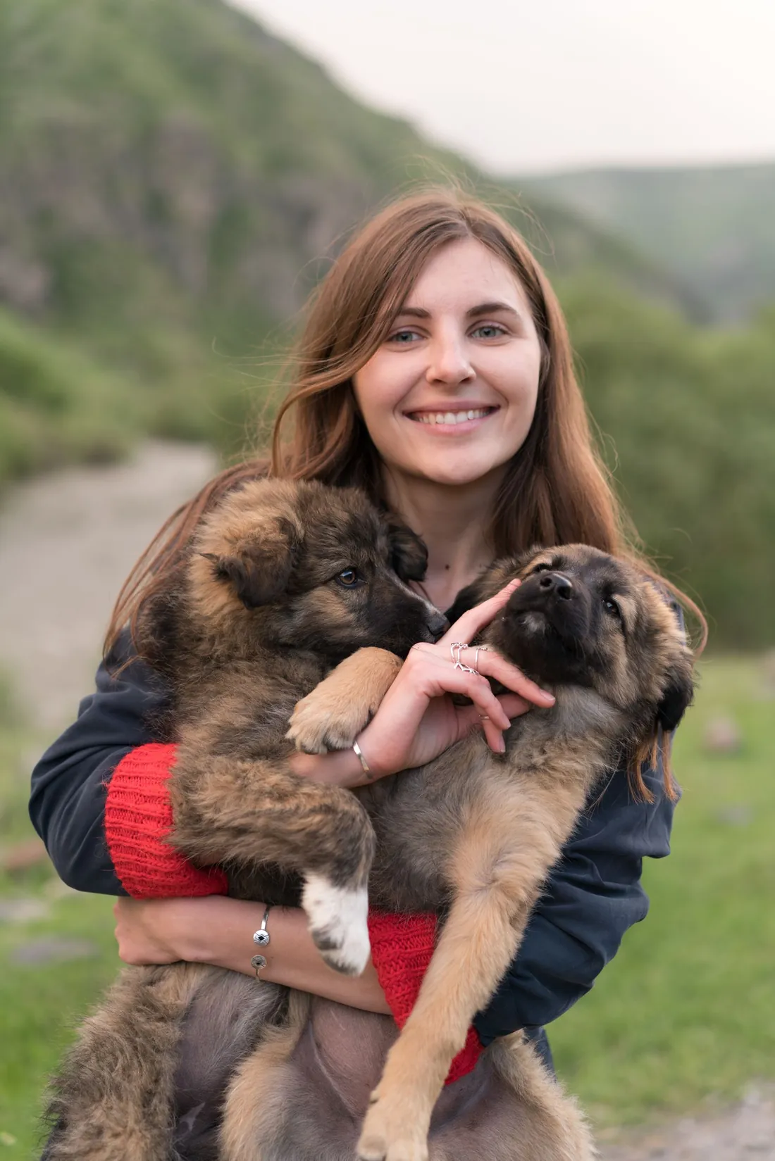 Une jeune femme sourit en tenant deux chiots bergers dans les bras, en montagne.
